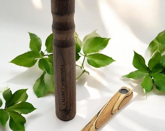 May include: A wooden smoking pipe with a brown and black wood grain pattern. The pipe is engraved with the words "Calumet Spiritpipes". The pipe is sitting on a white surface with green leaves in the background.