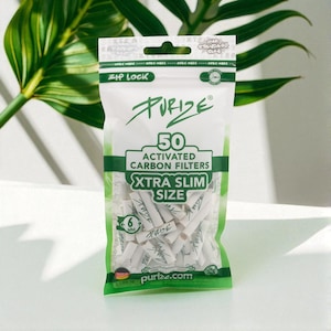 Peut inclure: Un paquet blanc et vert de filtres à charbon actif Purize. Le paquet indique "50 Filtres à charbon actif Xtra Slim Size" et "6 mm". Le paquet est scellé avec une fermeture éclair et contient de nombreux filtres blancs.