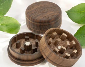 May include: A wooden grinder with a magnetic lid. The grinder has a dark brown wood finish and features a circular design with a central hole. The lid has the text "GRINDER BY CALMNESS EST 2015" engraved on it.