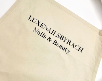 Könnte beinhalten: Eine beige Canvas-Schürze mit dem aufgedruckten Text "LUXENAILSBYRACH Nails & Beauty" in Schwarz.