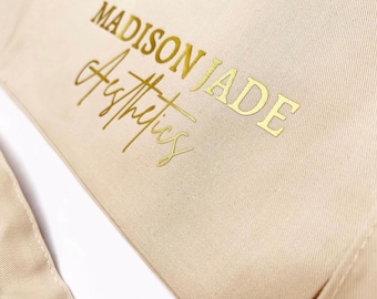 Könnte beinhalten: Beige Canvas-Tragetasche mit dem goldenen Text "MADISON JADE Aesthetics" auf der Vorderseite. Die Tasche hat ein schlichtes Design mit einem Riemen und wird wahrscheinlich zum Tragen von Gegenständen verwendet.
