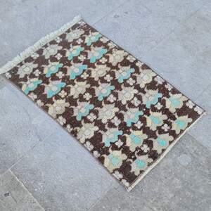 May include: A small brown rug with a floral pattern in shades of blue, white, and beige. The rug has a fringe border.