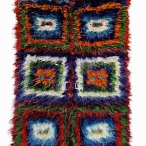 May include: A colourful, rectangular rug with a geometric pattern. The rug displays a repeating square design in red, orange, blue, green, and white. The top edge has white tassels. The rug is made from a shaggy, textured material, and would suit a living room or bedroom.