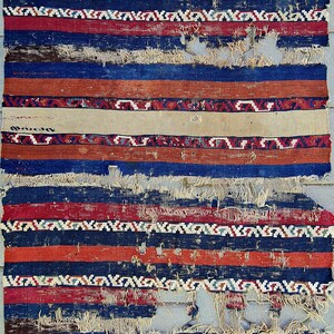 K&ouml;nnte beinhalten: Ein Vintage-Teppich mit einem gestreiften Muster in Rot-, Blau- und Wei&szlig;t&ouml;nen. Der Teppich ist abgenutzt und ausgefranst und zeigt Spuren von Alter und Gebrauch.