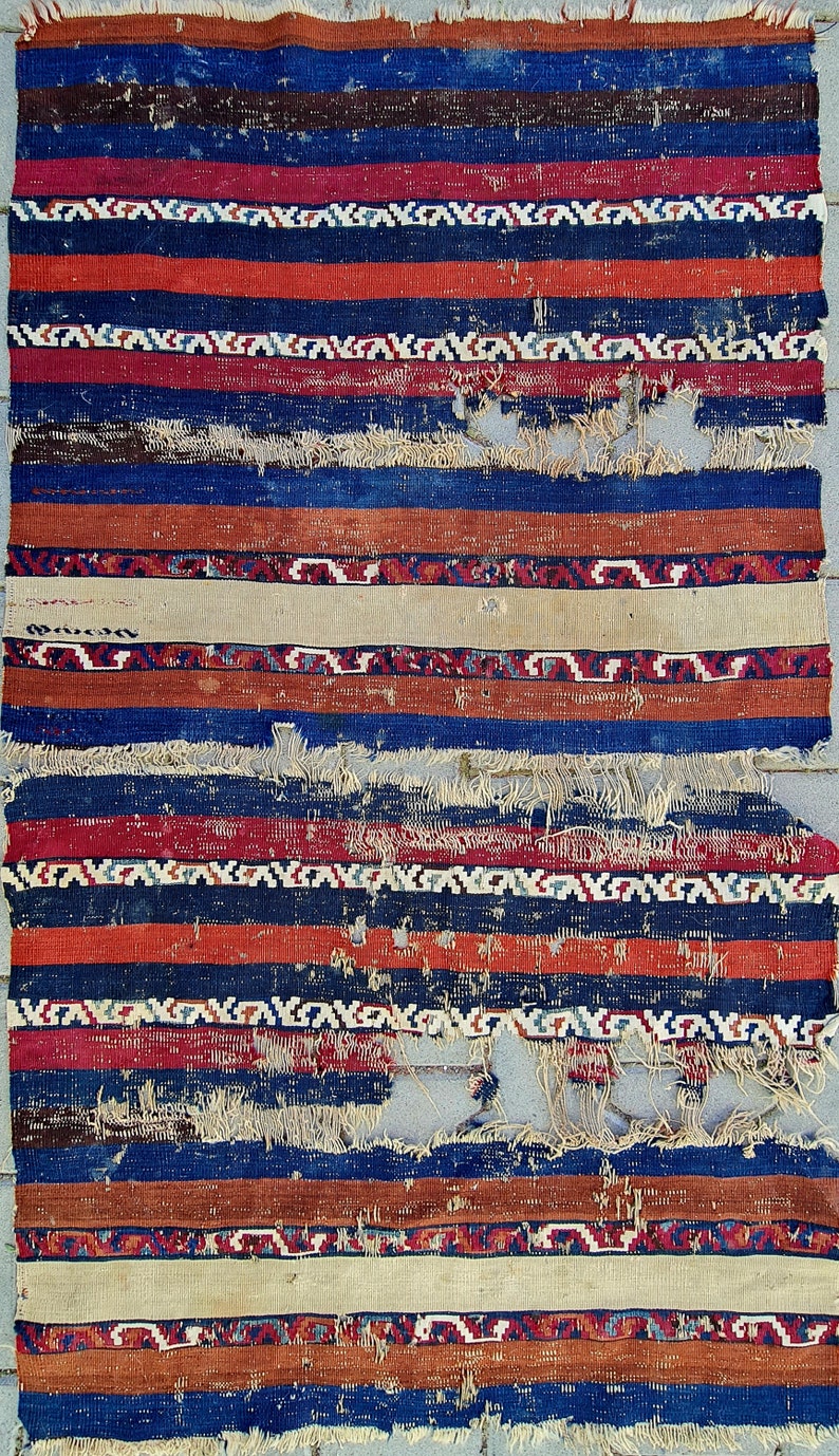 K&ouml;nnte beinhalten: Ein Vintage-Teppich mit einem gestreiften Muster in Rot-, Blau- und Wei&szlig;t&ouml;nen. Der Teppich ist abgenutzt und ausgefranst und zeigt Spuren von Alter und Gebrauch.
