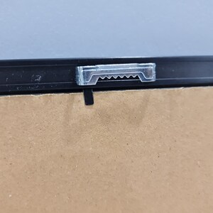 May include: A close-up of a black picture frame with a clear plastic hanging hook attached to the top.