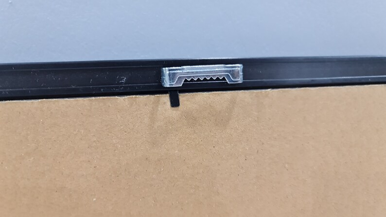 May include: A close-up of a black picture frame with a clear plastic hanging hook attached to the top.