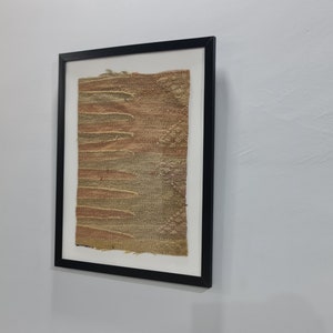 K&ouml;nnte beinhalten: Ein gerahmtes Kunstwerk mit einem strukturierten, gewebten Stoff in Braun- und Gr&uuml;nt&ouml;nen. Der Stoff hat eine raue, organische Textur und scheint handgewebt zu sein.