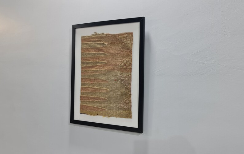 K&ouml;nnte beinhalten: Ein gerahmtes Kunstwerk mit einem strukturierten, gewebten Stoff in Braun- und Gr&uuml;nt&ouml;nen. Der Stoff hat eine raue, organische Textur und scheint handgewebt zu sein.