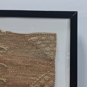 K&ouml;nnte beinhalten: Ein gerahmtes St&uuml;ck gewebten Stoffes mit einem braunen und beigefarbenen Muster. Der Stoff hat eine raue Textur und wirkt alt.