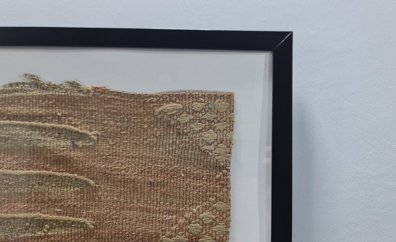 K&ouml;nnte beinhalten: Ein gerahmtes St&uuml;ck gewebten Stoffes mit einem braunen und beigefarbenen Muster. Der Stoff hat eine raue Textur und wirkt alt.