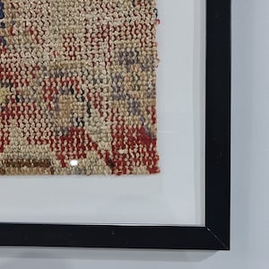 May include: A framed piece of woven fabric with a red, brown, and beige pattern. The fabric is in a black frame with a white mat.