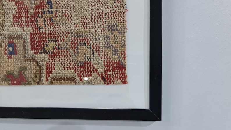 May include: A framed piece of woven fabric with a red, brown, and beige pattern. The fabric is in a black frame with a white mat.