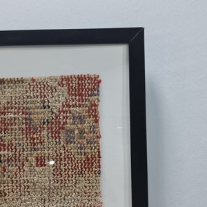 May include: A framed piece of woven fabric with a red, blue, and beige pattern. The fabric is displayed in a black frame with a white mat.