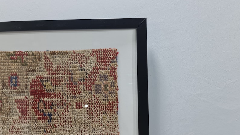 May include: A framed piece of woven fabric with a red, blue, and beige pattern. The fabric is displayed in a black frame with a white mat.
