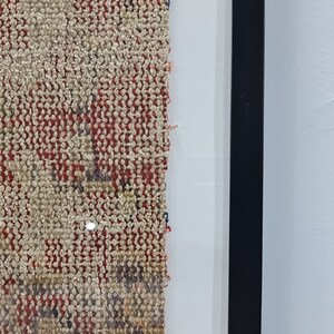 May include: Close-up of a framed piece of woven fabric with a red, white, and blue pattern. The fabric is frayed on the edges and has a textured surface.