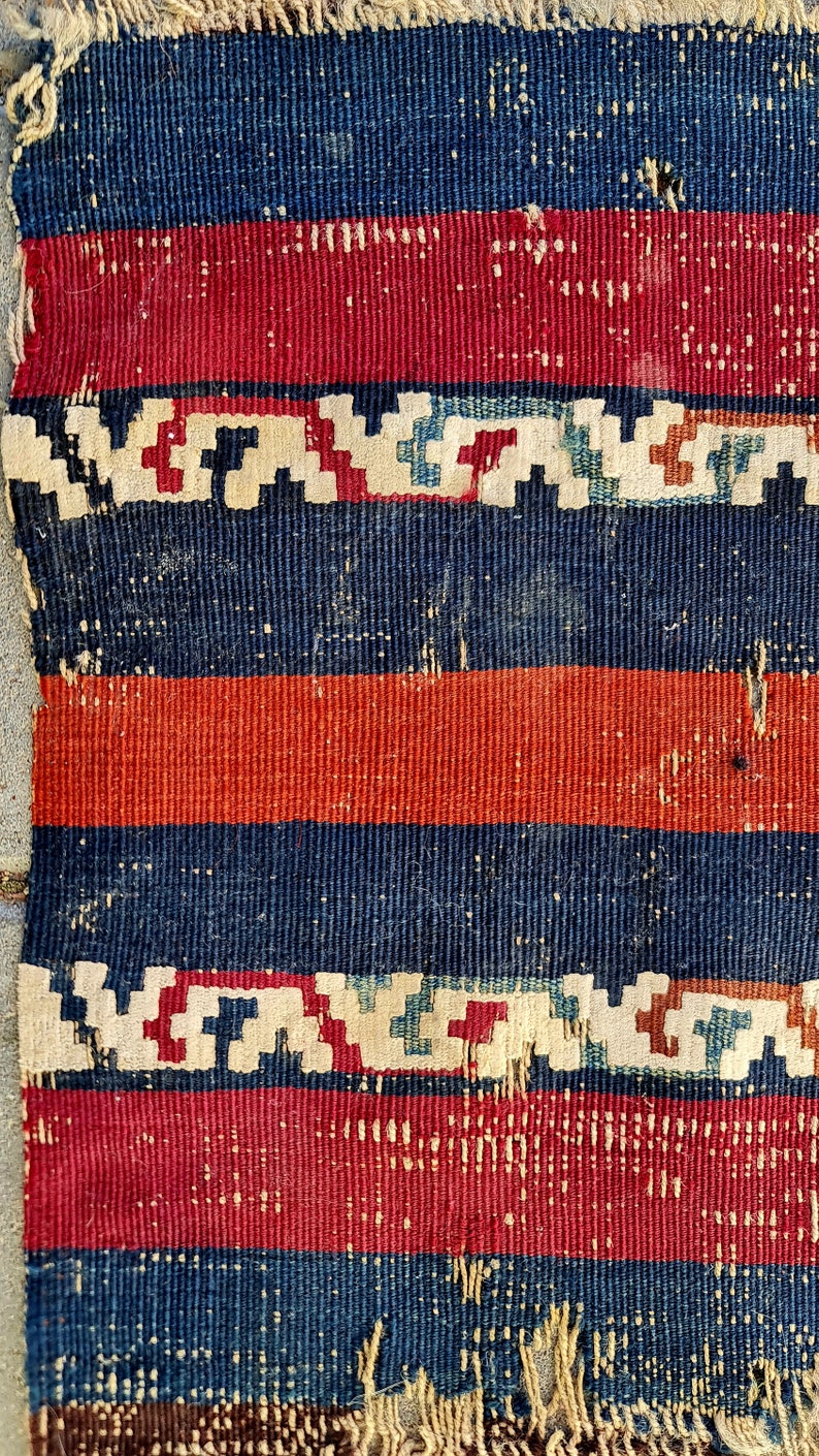 K&ouml;nnte beinhalten: Nahaufnahme eines gewebten Teppichs mit einem sich wiederholenden Muster aus roten, blauen und wei&szlig;en Streifen. Der Teppich hat einen ausgefransten Rand und weist Gebrauchsspuren auf.