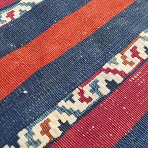 K&ouml;nnte beinhalten: Nahaufnahme eines gewebten Teppichs mit roten, blauen und wei&szlig;en Streifen. Der Teppich hat ein geometrisches Muster in den wei&szlig;en Streifen.