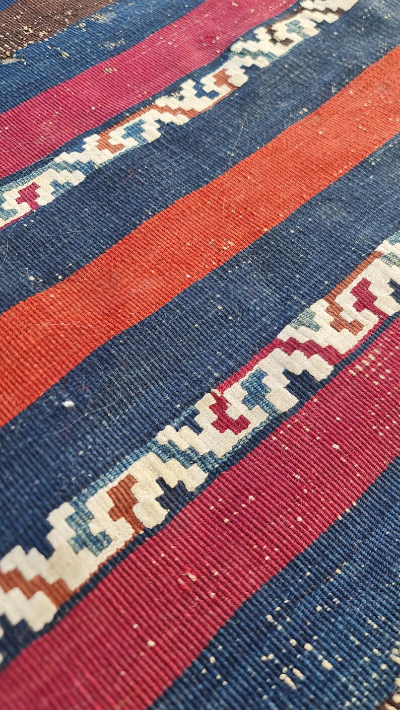 K&ouml;nnte beinhalten: Nahaufnahme eines gewebten Teppichs mit roten, blauen und wei&szlig;en Streifen. Der Teppich hat ein geometrisches Muster in den wei&szlig;en Streifen.