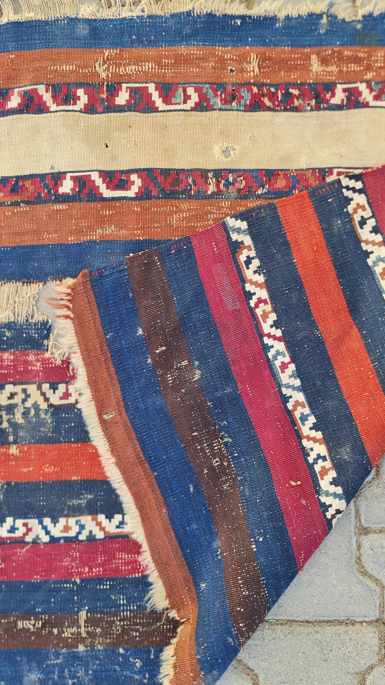 K&ouml;nnte beinhalten: Ein Vintage-Streifen-Teppich mit ausgefransten R&auml;ndern. Der Teppich ist aus Wolle und hat eine Vielzahl von Farben, darunter Rot, Blau, Braun und Wei&szlig;.