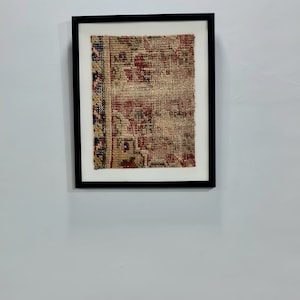 May include: A framed piece of antique rug with a red, blue, and beige pattern. The rug is in a black frame with a white mat.