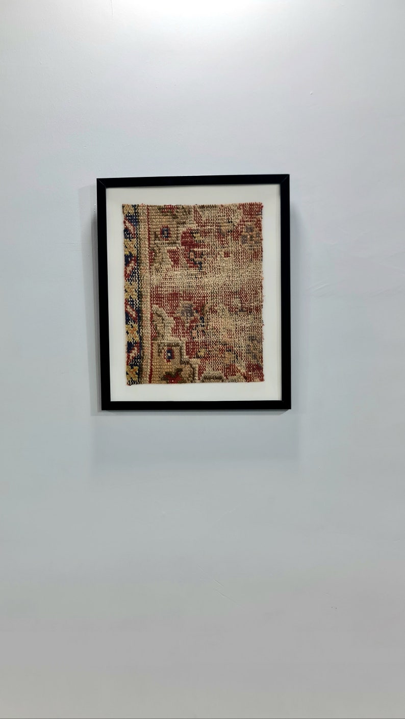 May include: A framed piece of antique rug with a red, blue, and beige pattern. The rug is in a black frame with a white mat.