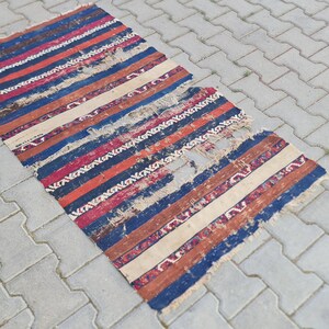 K&ouml;nnte beinhalten: Ein abgenutzter, gestreifter Teppich mit einem Loch in der Mitte. Der Teppich ist aus Wolle und hat verschiedene Farben, darunter Rot, Blau, Wei&szlig; und Braun.