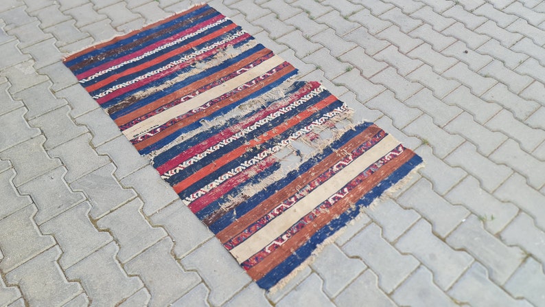 K&ouml;nnte beinhalten: Ein abgenutzter, gestreifter Teppich mit einem Loch in der Mitte. Der Teppich ist aus Wolle und hat verschiedene Farben, darunter Rot, Blau, Wei&szlig; und Braun.