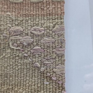 K&ouml;nnte beinhalten: Nahaufnahme einer gewebten Wandteppich mit einem rosa und beigefarbenen Muster. Der Wandteppich ist in einem schwarzen Rahmen gerahmt.