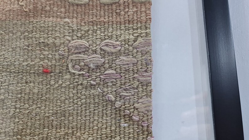 K&ouml;nnte beinhalten: Nahaufnahme einer gewebten Wandteppich mit einem rosa und beigefarbenen Muster. Der Wandteppich ist in einem schwarzen Rahmen gerahmt.