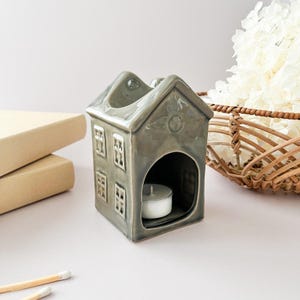 Peut inclure: Un brûleur d'huile en céramique grise en forme de maison avec une bougie chauffe-plat à l'intérieur. La maison a quatre fenêtres et un emblème décoratif sur le toit. Un panier de fleurs blanches et une pile de livres sont en arrière-plan.