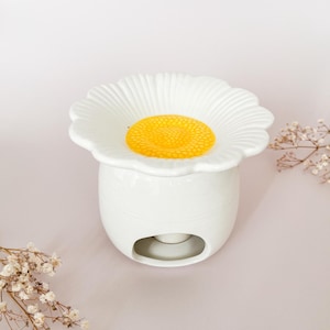 Peut inclure: Un chauffe-cire en céramique blanche en forme de fleur de marguerite. Le bol supérieur présente des pétales blancs rayonnant autour d'un centre jaune vif texturé. La base est un récipient cylindrique blanc lisse avec une grande ouverture arquée.