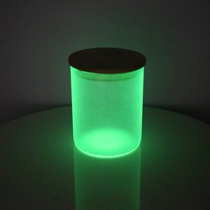 May include: A frosted glass jar with a wooden lid glows green in the dark. The jar is sitting on a white surface.