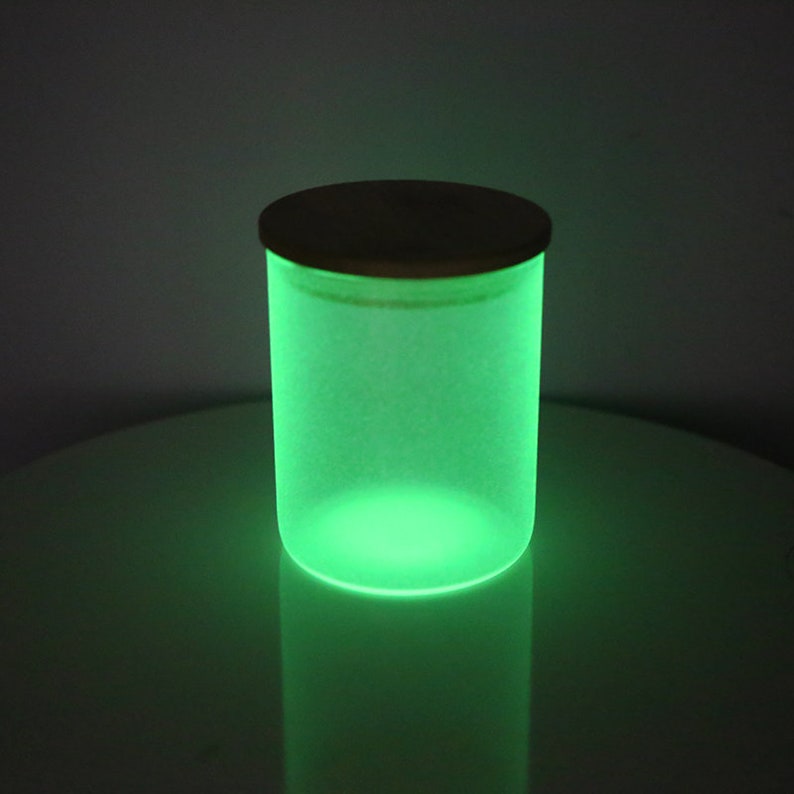 May include: A frosted glass jar with a wooden lid glows green in the dark. The jar is sitting on a white surface.