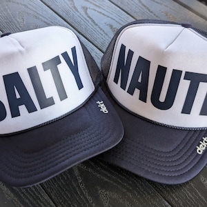 May include: Two navy blue and white trucker hats. One hat has the word "SALTY" printed in bold, dark gray letters. The other hat has the word "NAUTI" printed in bold, dark gray letters. Both hats have a mesh back and a curved brim.