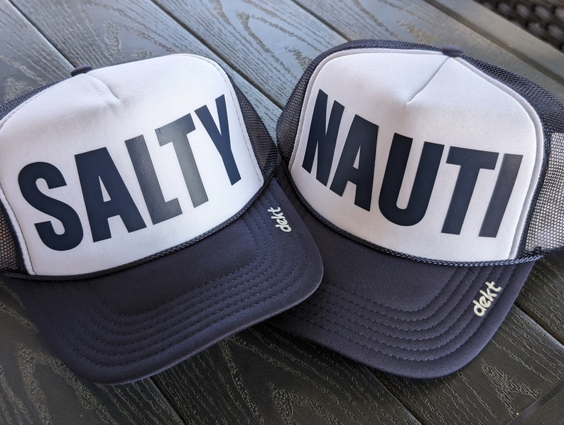 May include: Two navy blue and white trucker hats. One hat has the word "SALTY" printed in bold, dark gray letters. The other hat has the word "NAUTI" printed in bold, dark gray letters. Both hats have a mesh back and a curved brim.