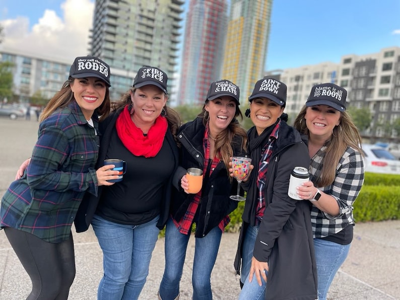 Peut inclure: Cinq femmes portant des casquettes de baseball noires avec des inscriptions diff&eacute;rentes. Les casquettes disent "They Still Call the Rodeo", "Fire & Ice", "Cowboy & Hats", "Ain't Goin Down", et "Blame it all on my Roots".
