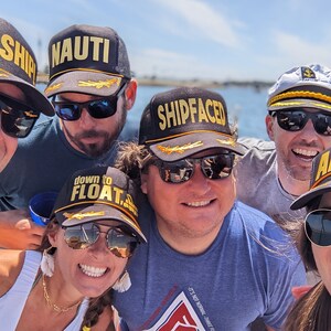 May include: A group of friends wearing black and grey baseball caps with gold lettering. The caps have different phrases on them, including "Oh, Ship", "Nauti", "Shipfaced", "Down to Float", and "Ahoy".