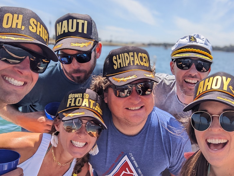 May include: A group of friends wearing black and grey baseball caps with gold lettering. The caps have different phrases on them, including "Oh, Ship", "Nauti", "Shipfaced", "Down to Float", and "Ahoy".