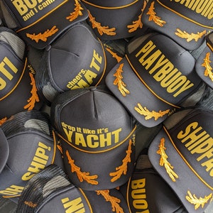 May include: A close-up of a pile of black trucker hats with gold embroidery. The hats feature various nautical-themed phrases like "Ahoy", "Playbuoy", "Drop it like it's Yacht", and "I like big boats and I cannot lie".