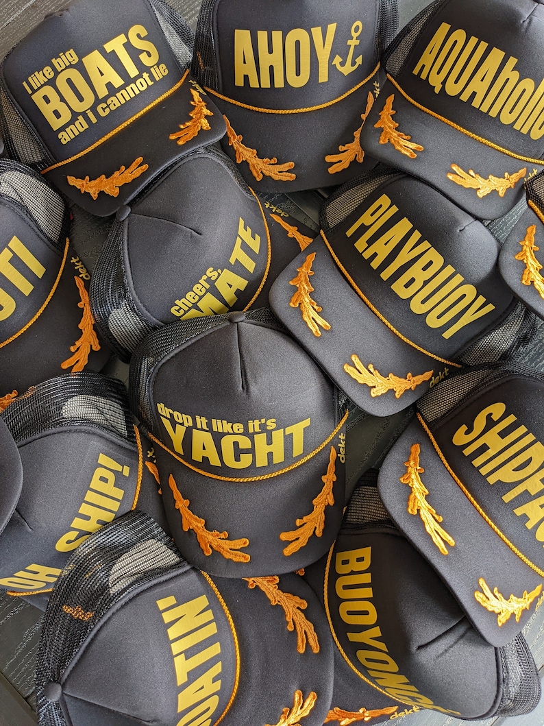 May include: A close-up of a pile of black trucker hats with gold embroidery. The hats feature various nautical-themed phrases like "Ahoy", "Playbuoy", "Drop it like it's Yacht", and "I like big boats and I cannot lie".