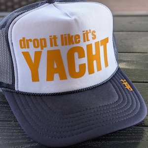 May include: A trucker hat with a white front panel and a navy blue brim and mesh back. The front panel features the text "drop it like it's YACHT" in orange. The hat has a curved brim and a rope detail along the edge.