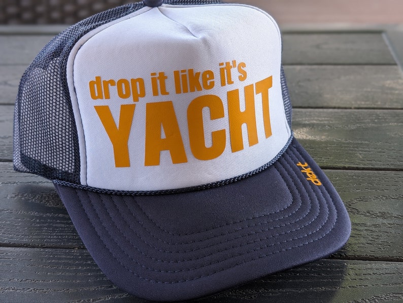 May include: A trucker hat with a white front panel and a navy blue brim and mesh back. The front panel features the text "drop it like it's YACHT" in orange. The hat has a curved brim and a rope detail along the edge.