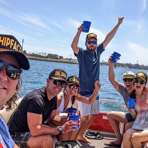 May include: A group of friends on a boat wearing hats with the words "SHIPFACE", "OH. SHIP", "FLOAT LIKE", "NAUTI", and "BOYZ" on them. They are holding blue cups and smiling.