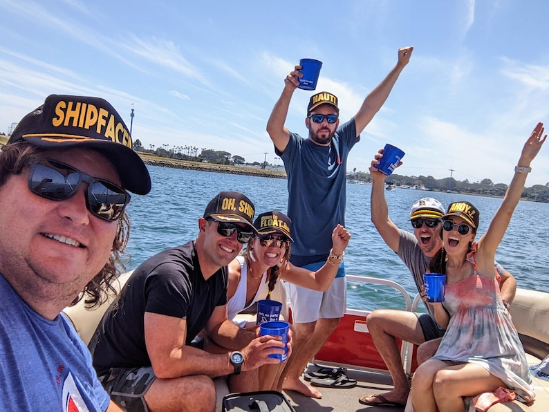 May include: A group of friends on a boat wearing hats with the words "SHIPFACE", "OH. SHIP", "FLOAT LIKE", "NAUTI", and "BOYZ" on them. They are holding blue cups and smiling.