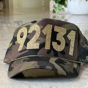 Könnte beinhalten: Eine Camouflage-Trucker-Kappe mit der goldenen Glitzerzahl "92131" auf der Vorderseite.