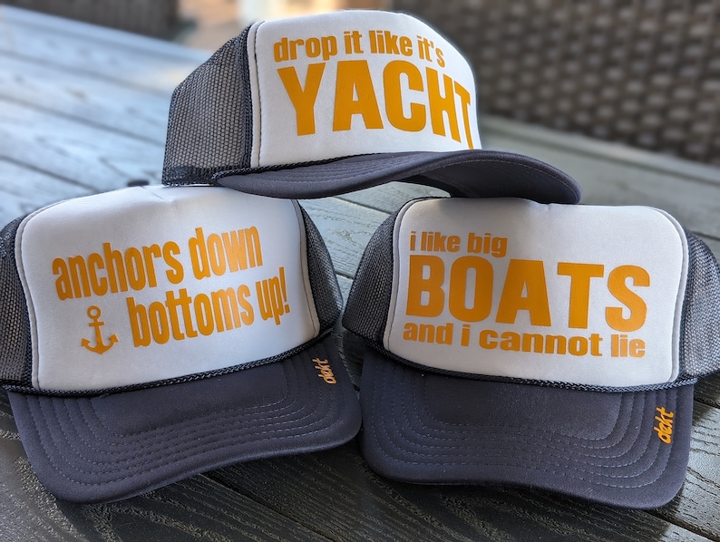 May include: Three trucker hats with white fronts and dark gray brims. The hats have mesh backs and feature orange text: "drop it like it's YACHT", "anchors down bottoms up!", and "i like big BOATS and i cannot lie".