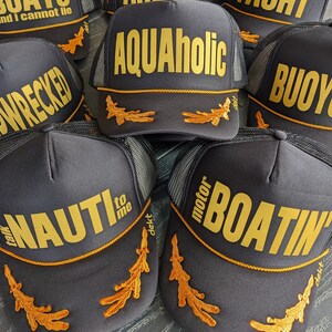 May include: Five black trucker hats with gold embroidered text and designs. The hats feature the words "Shipwrecked", "Aquaholic", "Buoyonce", "Just Nauti to me", and "Motor Boatin'" with a gold embroidered seahorse design on each hat.