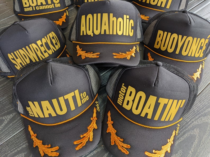May include: Five black trucker hats with gold embroidered text and designs. The hats feature the words "Shipwrecked", "Aquaholic", "Buoyonce", "Just Nauti to me", and "Motor Boatin'" with a gold embroidered seahorse design on each hat.