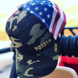 May include: A camouflage baseball cap with a mesh back featuring an American flag design. The cap has gold glitter numbers "921314" and the word "dekt" on the front.